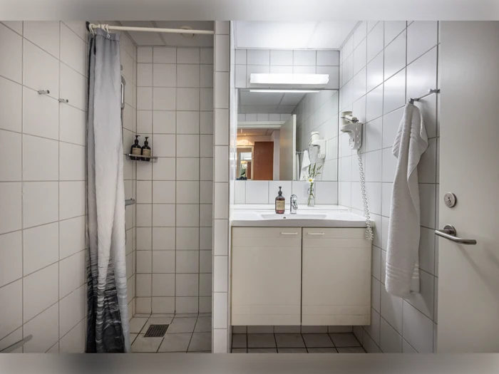 Et moderne badeværelse med hvide flisevægge, en åben bruser med forhæng, en håndvask med spejl og et håndklæde, der hænger på en krog på døren.