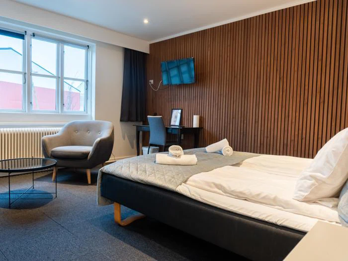 Moderne hotelværelse med dobbeltseng, lænestol, skrivebord, tv monteret på en accentvæg af træ, rundt glasbord og stort vindue, der lukker naturligt lys ind.