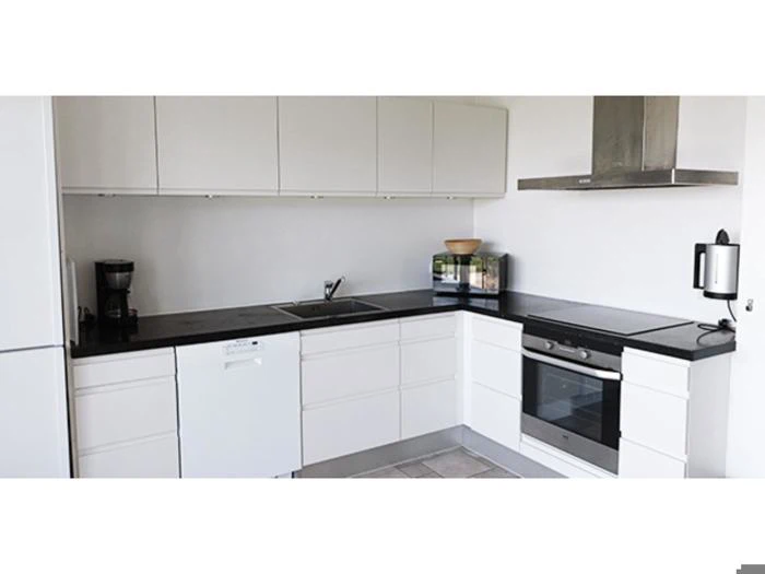 Moderne hvidt køkken med sorte bordplader, indbygget ovn, komfur, opvaskemaskine, vask, kaffemaskine, mikroovn og emhætte. Minimalistisk design med over- og underskabe.