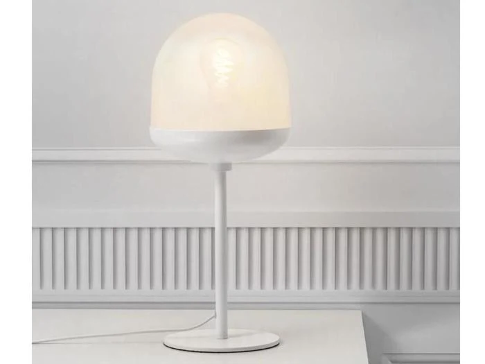 En moderne hvid bordlampe med en rund, matteret glasskærm står på en hvid flade mod en hvid panelvæg.
