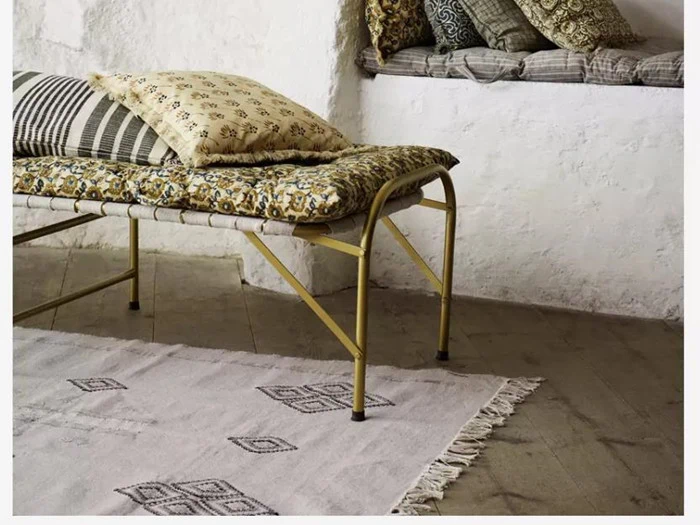 En metalindrammet daybed med mønstrede puder står på et struktureret tæppe ved siden af en hvidkalket væg med et indbygget vinduessæde og forskellige puder.