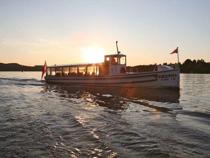 En hvid passagerbåd ved navn Viking sejler på en rolig sø ved solnedgang med rislende vand og træer i silhuet i baggrunden.