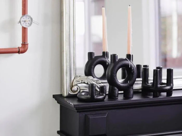 Sorte abstrakte lysestager med lyse stearinlys står på en sort kamin foran et sølvindrammet spejl; et vægur og en rød pibe ses til venstre.