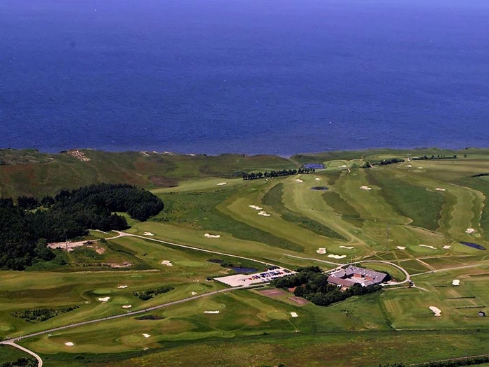 Luftfoto af en golfbane med sandbunkers, grønne fairways, et klubhus og en parkeringsplads, der ligger ved en kystlinje med blåt hav bagved.
