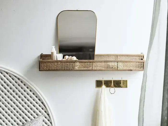En guldhylde med spejl, toiletsager og skaller over en guldknagerække med en hvid klud, monteret på en hvid væg ved siden af en seng med en hvid vævet hovedgærde.