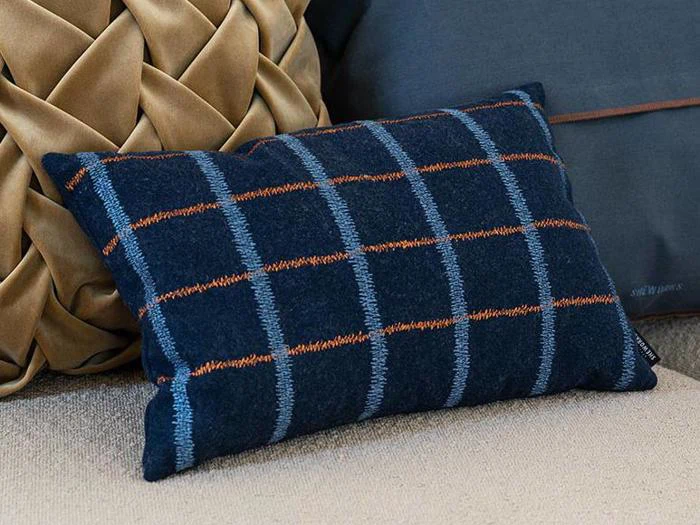 En rektangulær marineblå pude med orange og lyseblå tern hviler på en beige sofa ved siden af en solbrun pude med et struktureret, flettet design.