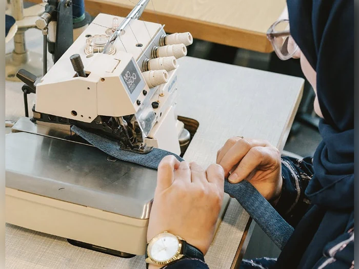 En person bruger en symaskine til at sy et stykke blåt stof, iført et ur og et mørkt ærme, med nærbillede af hænderne og maskinen.