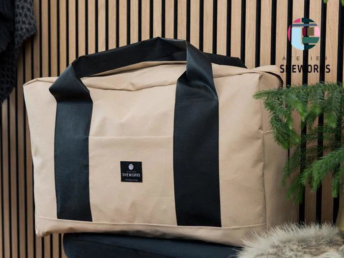 En beige duffelbag med sorte håndtag står på en mørk flade med en logopatch på forsiden. I baggrunden er der trælister og grønne planter, og et Atelier Sheworks-skilt er synligt.