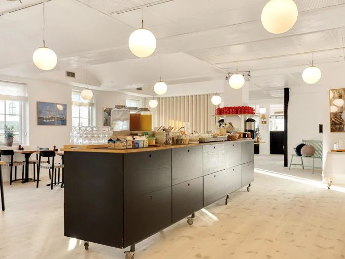 Moderne, lyst køkken med en stor sort ø på hjul, morgenmadsartikler, kuglependler og borde med stole i baggrunden.