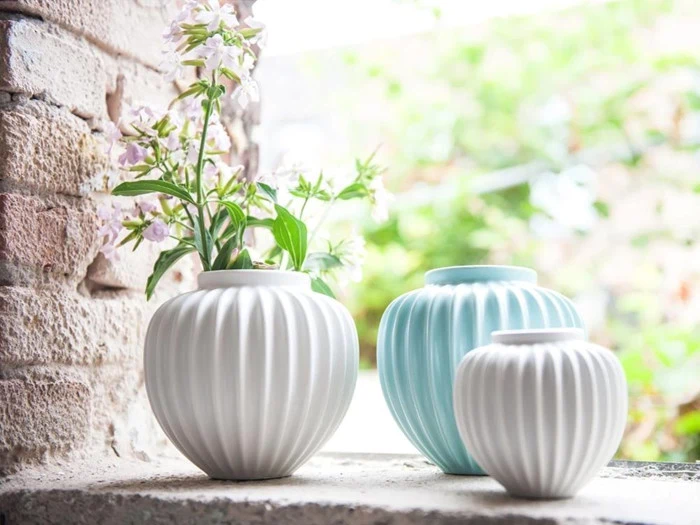 Tre riflede keramikvaser - en med blomster - står på en stenkant nær en murstensvæg med grønne planter og blødt sollys i baggrunden.