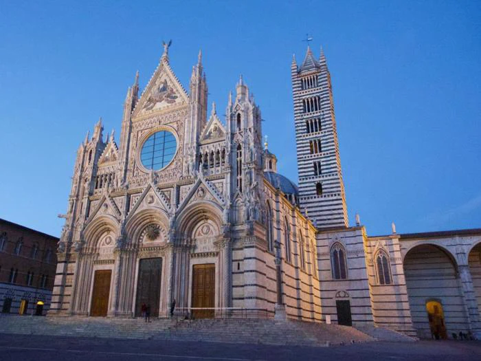 Sienas katedral i Italien med sin udsmykkede gotiske facade, rosenvindue og høje stribede klokketårn, oplyst mod en klar blå aftenhimmel.