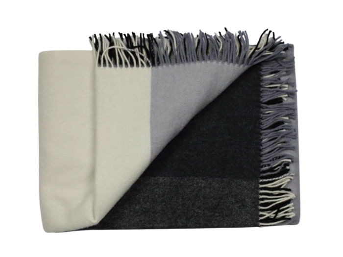 Et foldet tæppe med et farveblokdesign i creme, grå og sort og med frynsede kanter på den ene side.