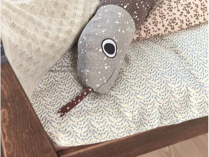 En plysslange med prikket mønster, store øjne og en tunge af stof ligger på et mønstret sengetæppe ved en sengeramme af træ.