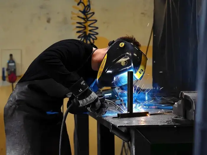 En person iført beskyttelsesudstyr og svejsehjelm svejser metalstykker sammen ved en arbejdsbænk, og det fyger med gnister.