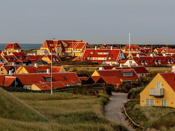 En kystby med gule huse og røde tage omgivet af græsklædte klitter under en overskyet himmel.