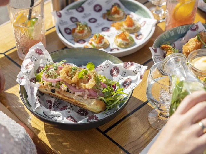 En sandwich med grønt og stegte løg på en tallerken, omgivet af forskellige appetitvækkere og drikkevarer på et solbeskinnet træbord.