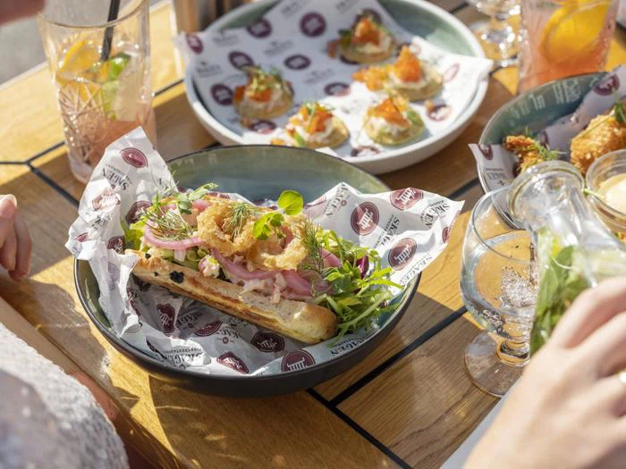 En sandwich med stegt pålæg, grønt og syltede løg ligger på mønstret papir i en skål, omgivet af tallerkener med appetitvækkere og drikkevarer på et træbord i sollys.