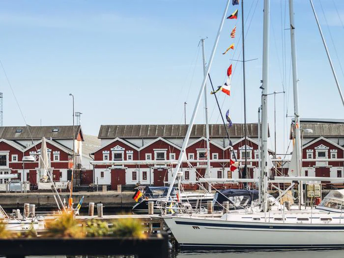 Hvide sejlbåde lægger til i en marina med røde lagerbygninger i baggrunden og farverige flag, der hænger fra en mast.