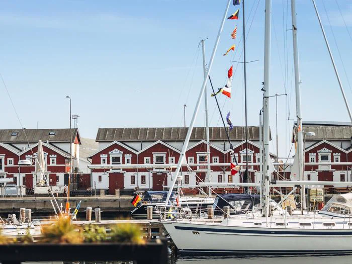 En marina med sejlbåde, der ligger foran røde træbygninger med hvide kanter under en klar blå himmel.