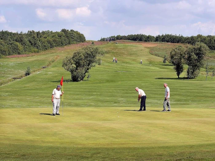 Tre mænd spiller golf på en grøn bane; en holder flagstangen, en gør klar til at putte, og en anden står i nærheden. Træer og bølgende bakker er synlige i baggrunden under en blå himmel.