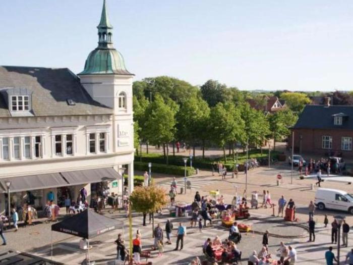 Et livligt bytorv med folk, der går og sidder ved udendørs borde, ved siden af en historisk bygning med et højt spir og omgivet af grønne træer på en solrig dag.