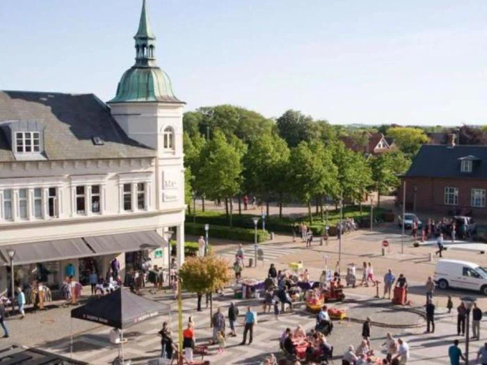 Et livligt bytorv med folk, der går og sidder ved udendørs borde, ved siden af en historisk bygning med et højt spir og omgivet af grønne træer på en solrig dag.