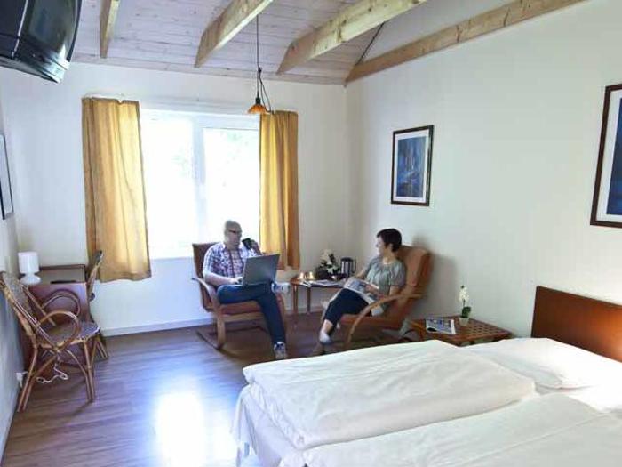 En mand og en kvinde sidder på stole omkring et lille bord i et lyst hotelværelse med trægulv, dobbeltseng og et stort vindue med gule gardiner.