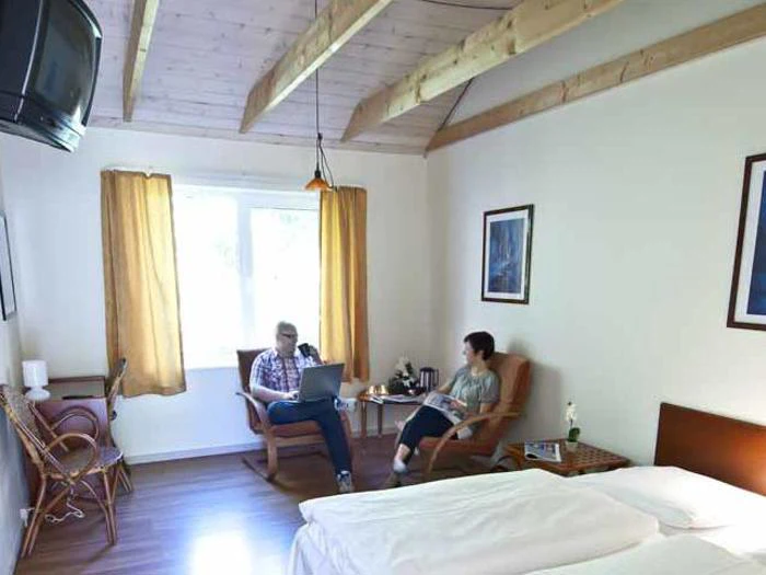 To personer sidder på stole i et lyst hotelværelse med træloft, et højt monteret tv og en seng med hvidt sengetøj. Den ene bruger en bærbar computer, mens den anden læser i en bog ved et vindue med gu