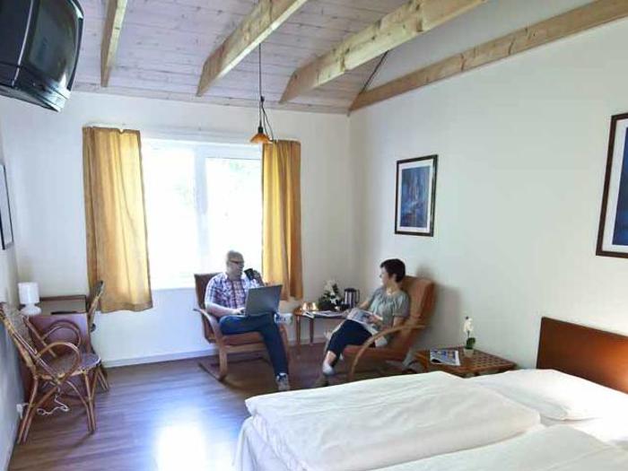 To personer sidder på stole i et lyst, hyggeligt hotelværelse med træbjælker i loftet, en seng, et vindue med gule gardiner og et væghængt tv.