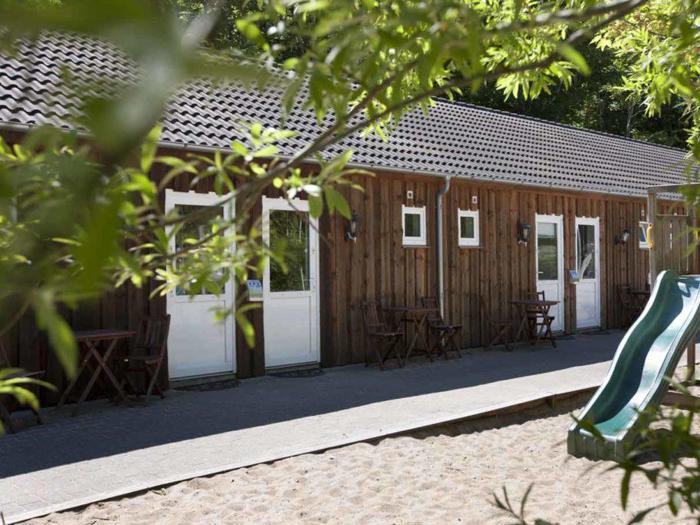 Træbygning i én etage med hvide døre og små borde udenfor, delvist indrammet af grønne grene, ved siden af en legeplads med rutsjebane og sand.