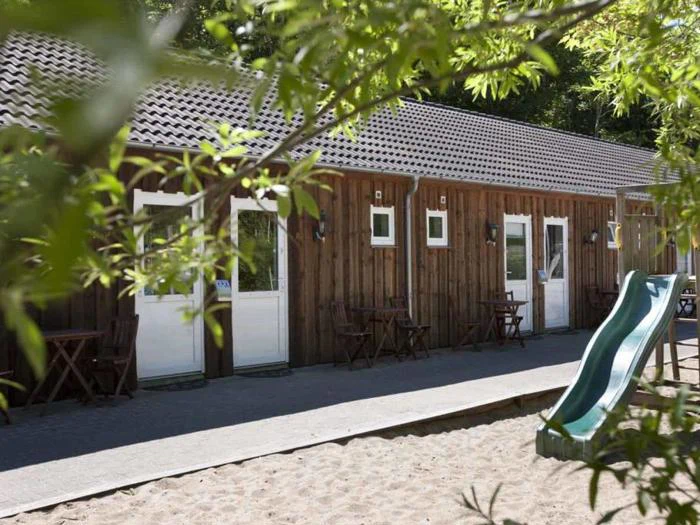 En træbygning med hvide døre og vinduer, udendørs borde og stole og en grøn rutsjebane på et sandet legepladsområde, delvist indrammet af grønne grene.