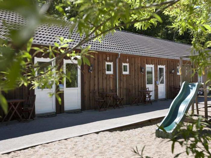 En træbygning med hvide døre og små vinduer, udendørs borde og en grøn legepladsrutsjebane i et sandområde, indrammet af grønne grene.