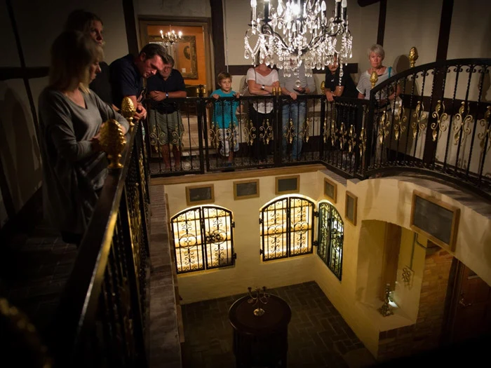 En gruppe mennesker står på en udsmykket balkon med udsigt til et vintage-interiør med lysekroner og store glasmosaikvinduer.