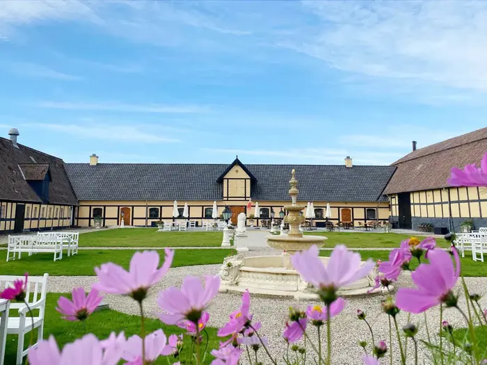 En gårdsplads med et centralt springvand af sten, omgivet af en stor gul bygning og blomstrende lyserøde blomster i forgrunden under en blå himmel.