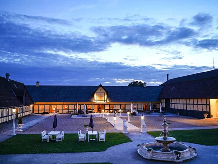 Et stort, elegant landsted med varmt lys, en central gårdsplads, udendørs siddepladser, statuer og et springvand i skumringen under en skyet blå himmel.