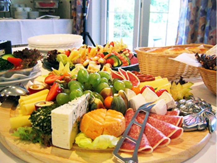 Et stort fad med forskellige oste, pålæg, vindruer, skiveskårne grøntsager og frisk brød er arrangeret på et bord, der er dækket op til buffet i et lyst lokale.