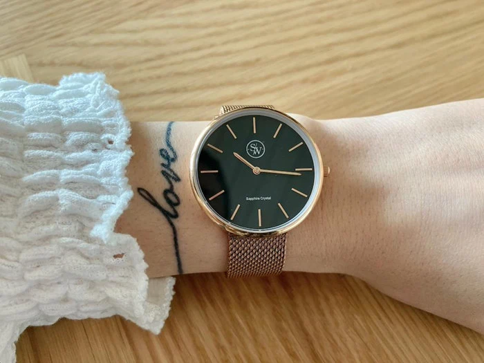 Der vises et håndled med et guldnetur og en sort urskive. Personen er iført et hvidt tekstureret ærme og har en kærlighedstatovering på håndleddet.