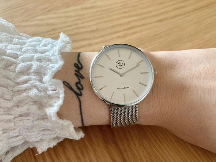 Et håndled iført et sølvfarvet mesh-ur og et hvidt tekstureret ærme med en kærlighedstatovering i kursiv på underarmen hviler på en træoverflade.