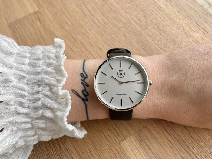 Et håndled med et sort læderur og en hvid urskive med ordet kærlighed tatoveret i kursiv på indersiden af håndleddet, der hviler på en lys træoverflade.