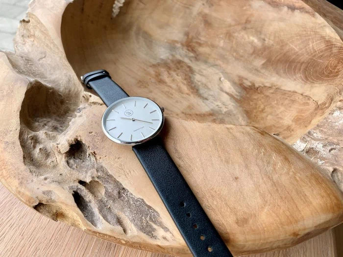 Et minimalistisk armbåndsur med sort læderrem hviler i en stor, udskåret træskål med naturlige teksturer.