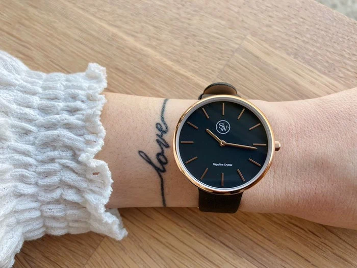 Et håndled med et ur i sort og guld og en kærlighedstatovering i kursiv, iført et hvidt tekstureret ærme, hviler på en træoverflade.