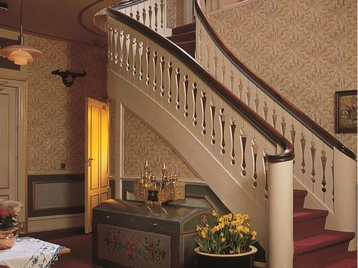 Buet trappe med rødt tæppe og dekorativt gelænder i et rum i vintagestil med udsmykket tapet, antikke møbler, en kiste med lysestager på toppen og en vase med gule blomster.