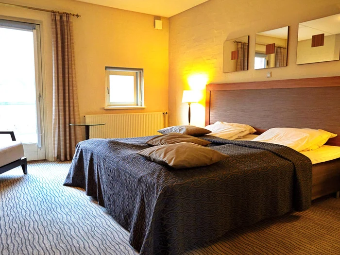 Et moderne hotelværelse med dobbeltseng, to puder, hovedgærde, spejlvæg, lampe, stol, glasbord og et stort vindue med gardiner, der lukker naturligt lys ind.