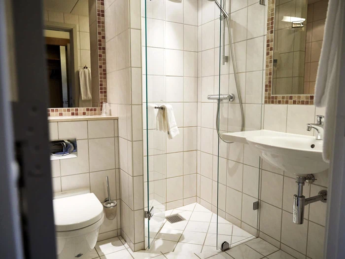 Et moderne badeværelse med hvide fliser, et væghængt toilet, en brusekabine af glas, en hvid håndvask og et spejl over håndvasken. Der hænger et håndklæde ved siden af bruseren.