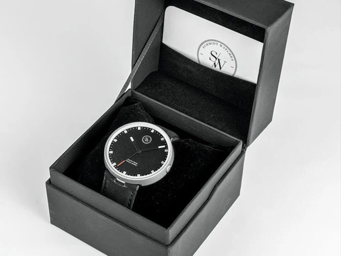 Et sort armbåndsur med rund urskive i en sort gaveæske ved siden af et mærkevarekort i æskens låg.