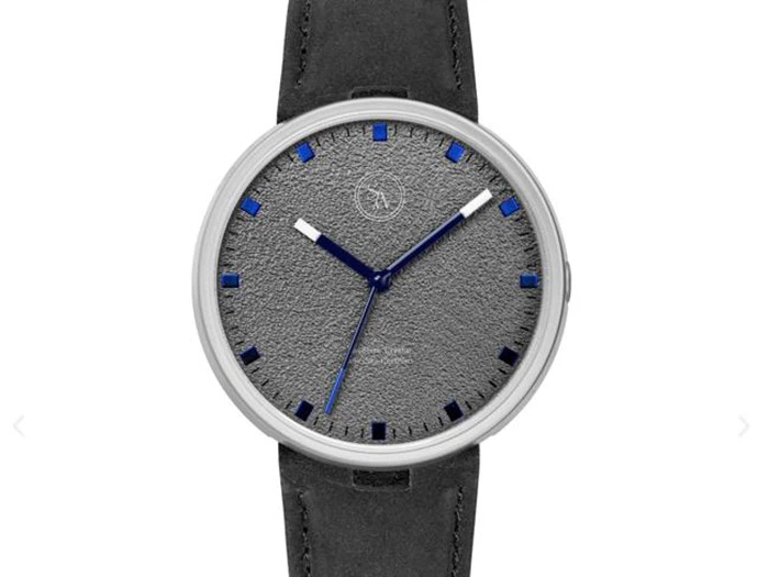 Et armbåndsur med en struktureret grå urskive, blå timemarkeringer, blå og sølvfarvede visere og en sort læderrem, vist på en hvid baggrund.