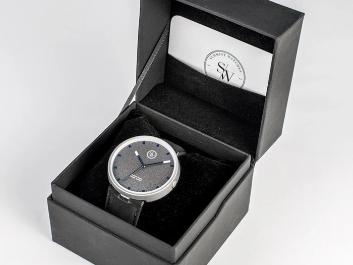 Et armbåndsur med sort rem og rund, sort urskive ligger i en åben, sort gaveæske med et visitkort i låget.