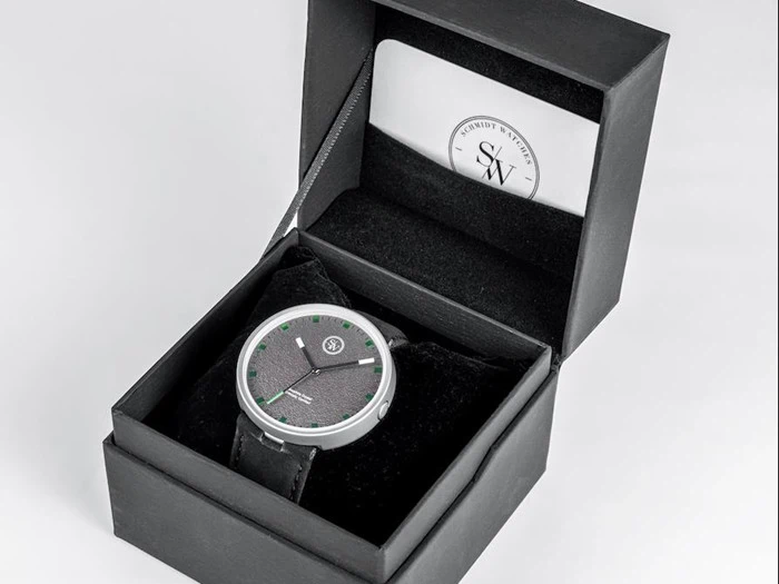Et sort armbåndsur med grønne timemarkeringer i en sort gaveæske, ledsaget af et kort med logoet SW på indersiden af låget.