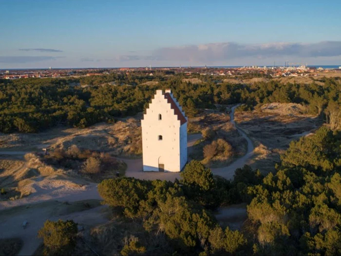 Et højt, hvidt kirketårn med gavle står alene blandt træer og sandede stier, og i det fjerne kan man se en by under en klar himmel.