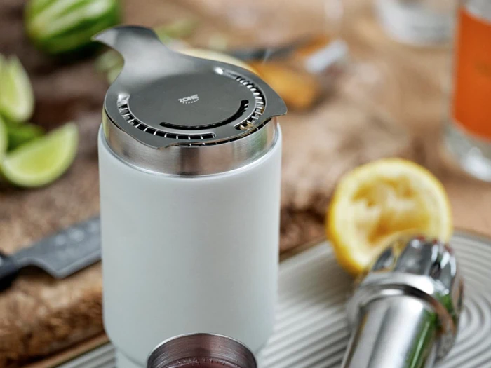 Et nærbillede af en cocktailsi af metal oven på en hvid shaker med en presset citron, en kniv og limebåde i baggrunden.